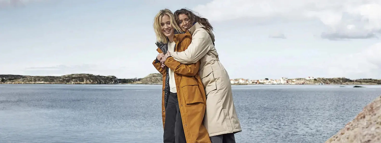 Two women embracing outdoors by water in stylish Didriksons country clothing.