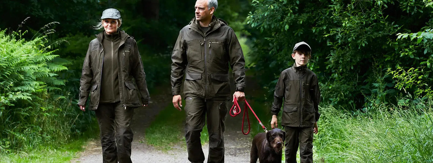 Three people walking a black dog on a path in the great outdoors, showcasing country clothing.