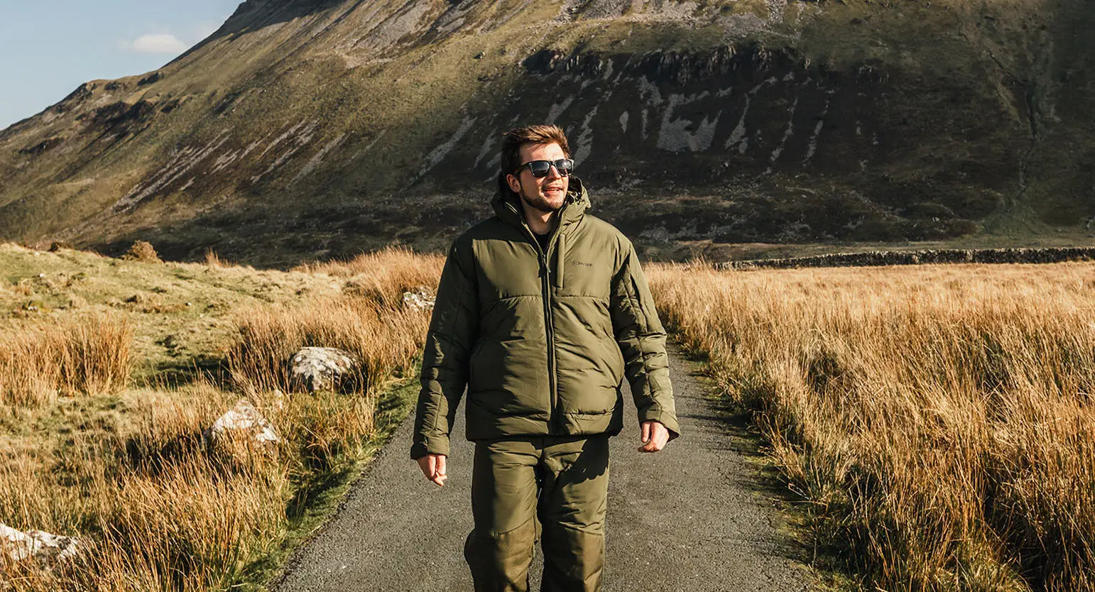 Man in a green jacket strolls through a grassy field, showcasing high quality outdoor gear.