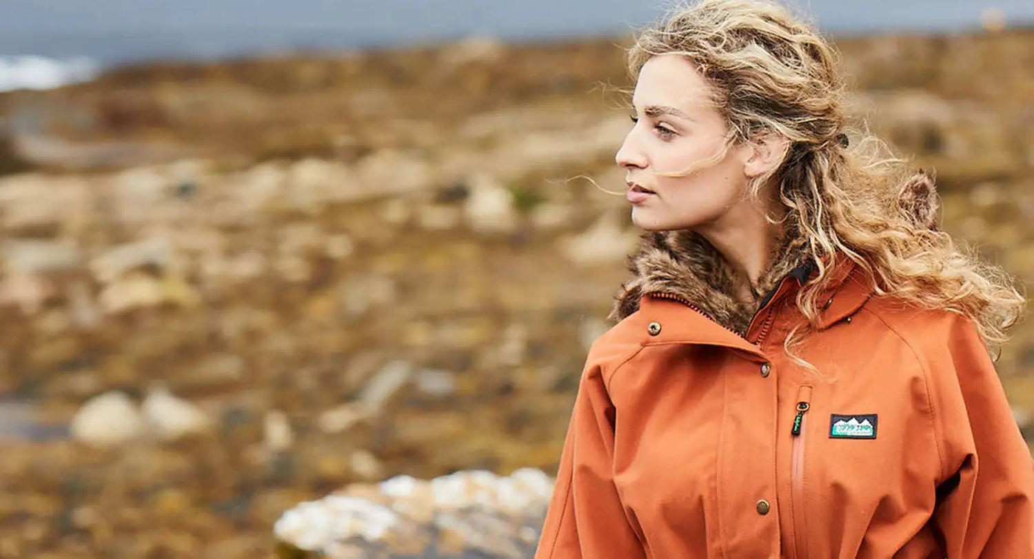 Woman with wavy blonde hair in orange Ridgeline jacket near a rugged mountain peak.