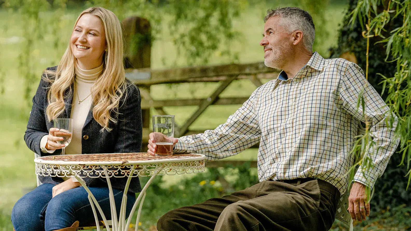 Two friends in tattersall shirts enjoying drinks at an outdoor table together.