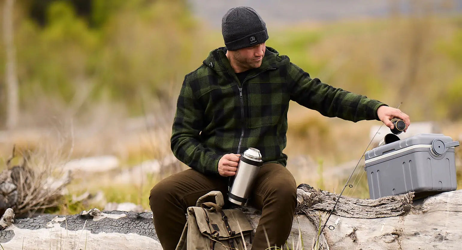 Man in plaid jacket and beanie enjoying the outdoors with a thermos and tackle box.