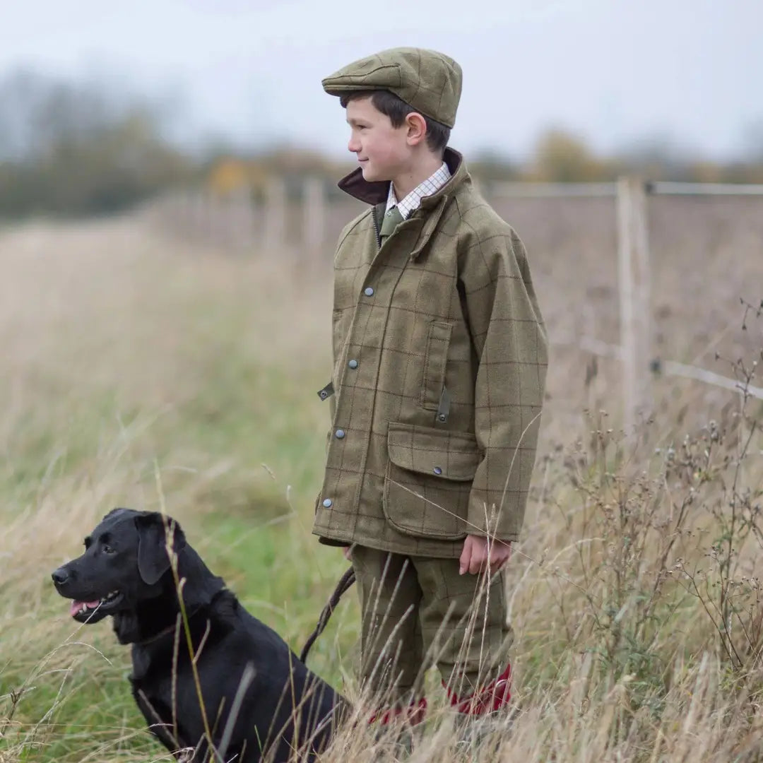 Young person in countryside attire with a black dog by a field, wearing Alan Paine Rutland cap