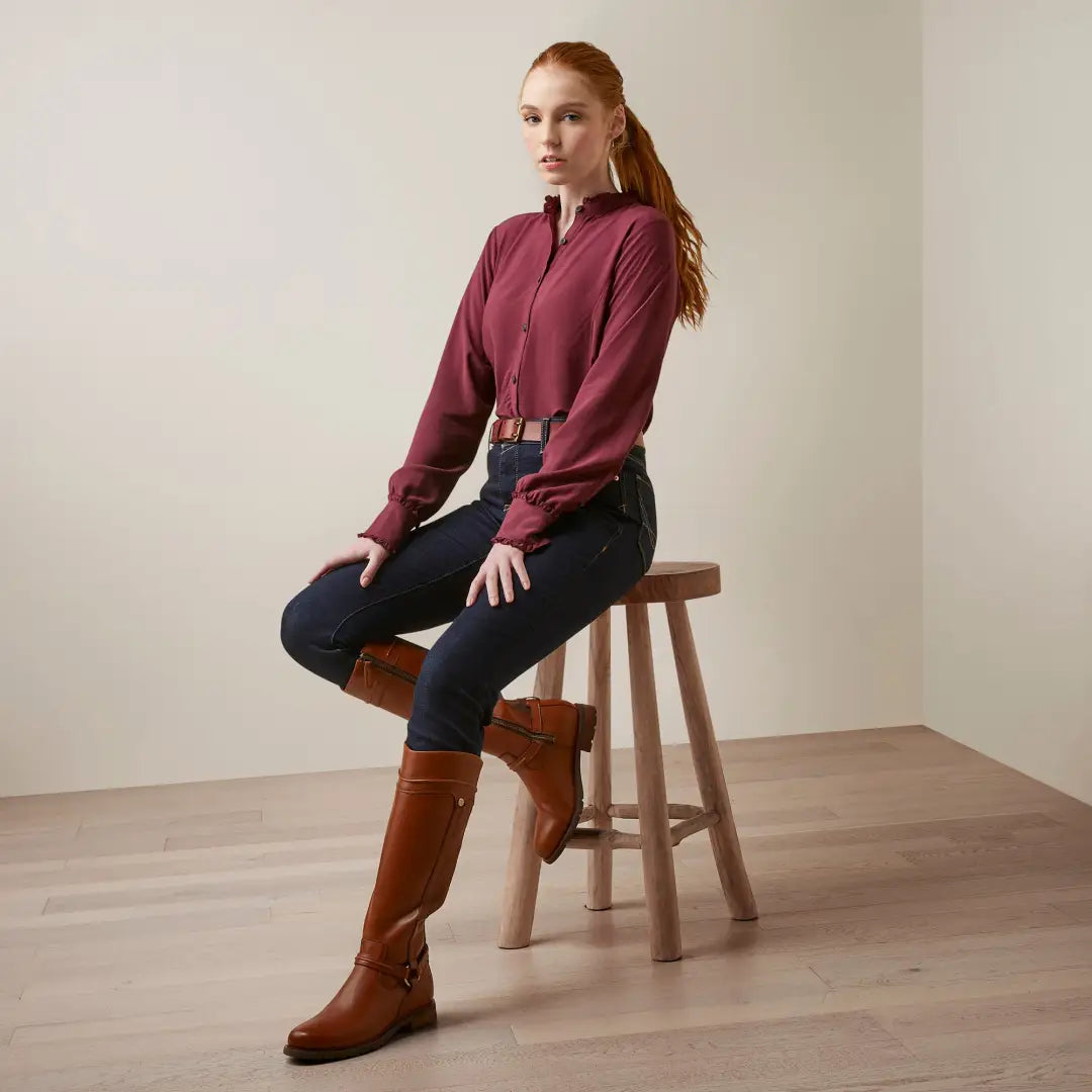 Woman in burgundy top and dark jeans showcases stylish country clothing on wooden stool