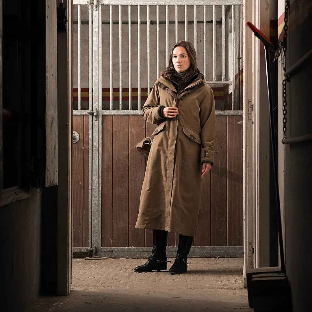 Woman in a beige Baleno Kensington coat by a horse stall, showcasing stylish outerwear