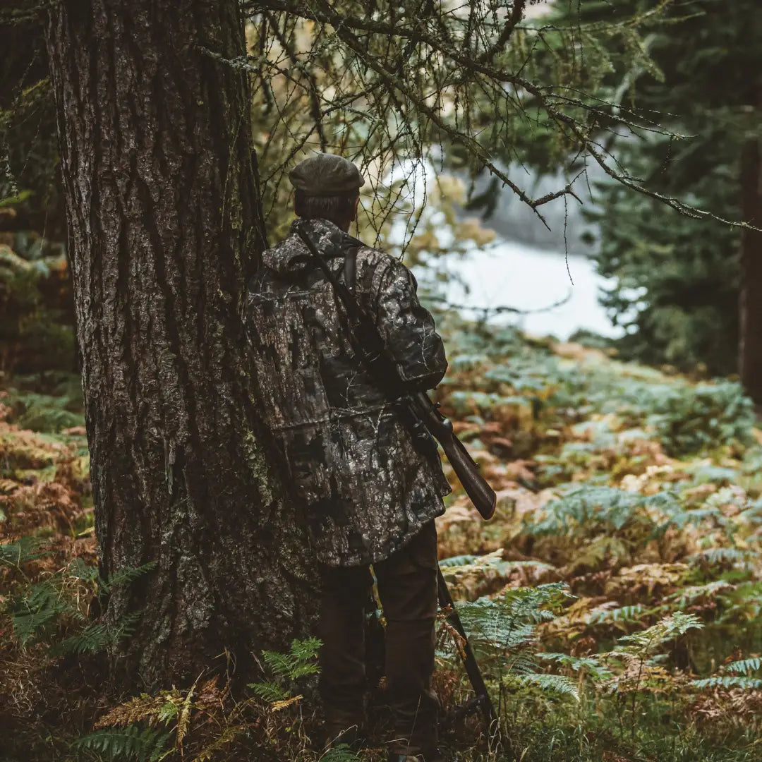 Camouflaged hunter by tree in forest wearing Deerhunter PRO Gamekeeper Hunting Jacket