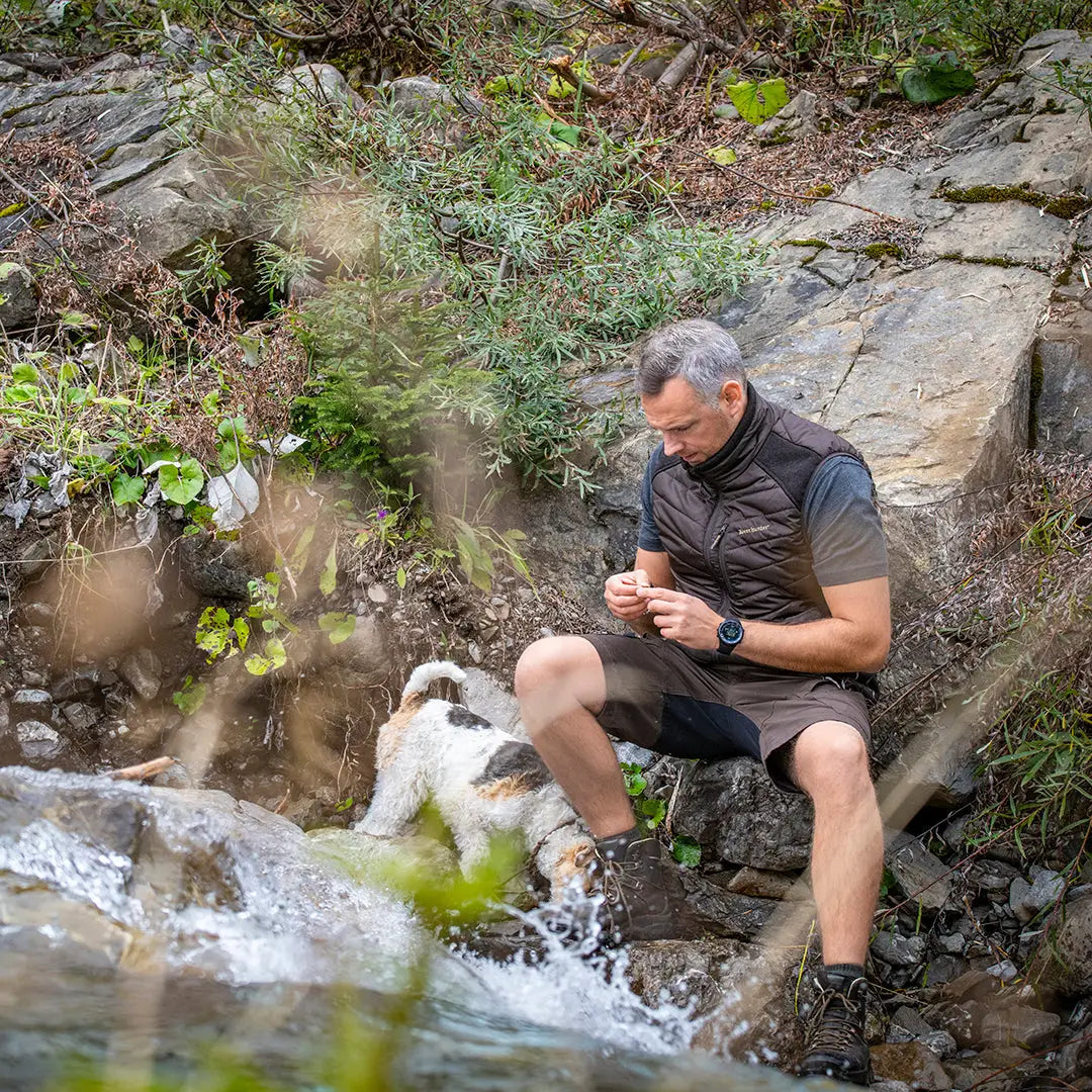 Man in Deerhunter Strike Shorts sitting by a stream examining something in hands