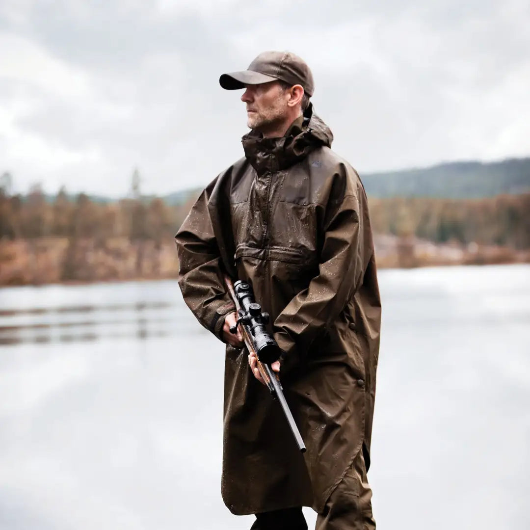 Man in a brown coat with a rifle by water wearing a Deerhunter Track Rain Anorak