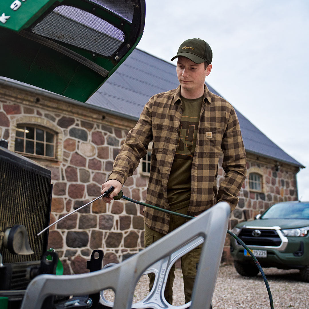 Man in plaid shirt using tractor while wearing Harkila Impact Cap for country clothing