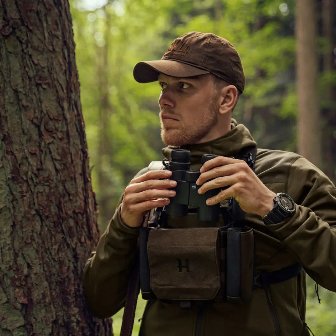 Man in a forest wearing Harkila Modi Cap while holding binoculars for spotting wildlife