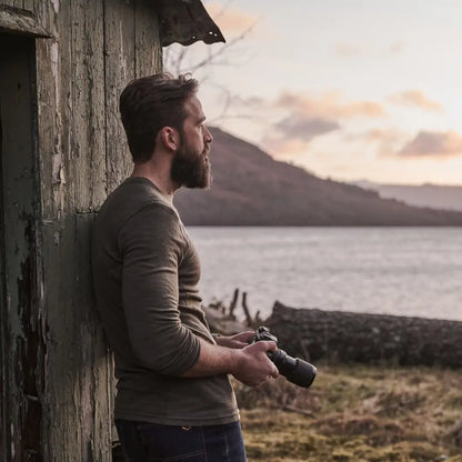 Bearded man in Merino wool crew neck top holding camera by wooden structure