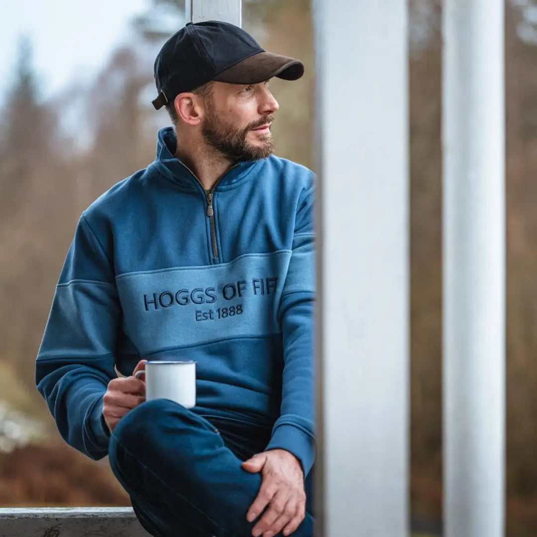 Man in blue Hoggs Of Fife quarter zip sweatshirt holding a mug, rocking a black cap