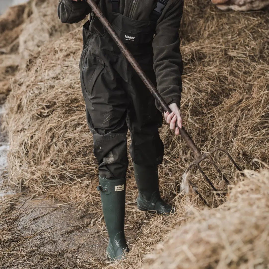 Person in dark outdoor gear and green boots holds a pitchfork beside hay, showcasing Fife Green King