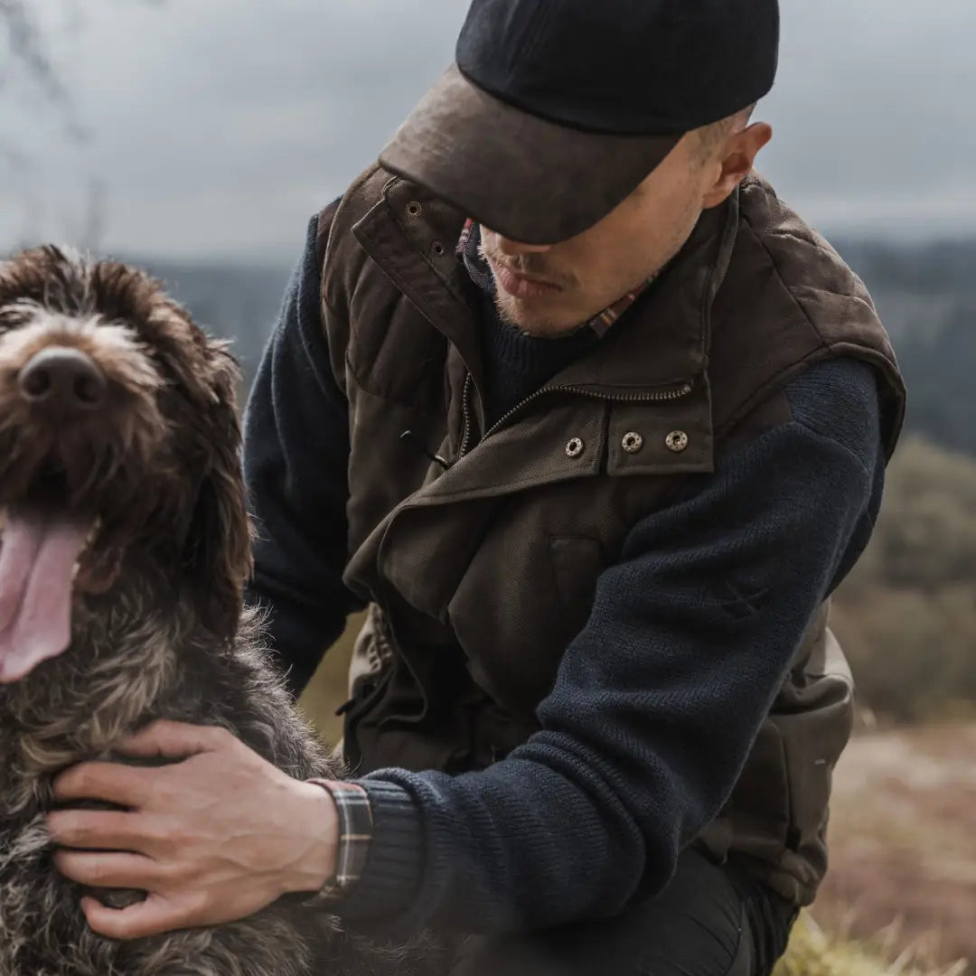 Man in a cap and jacket pets a shaggy brown dog while wearing a Kincraig Field Waistcoat