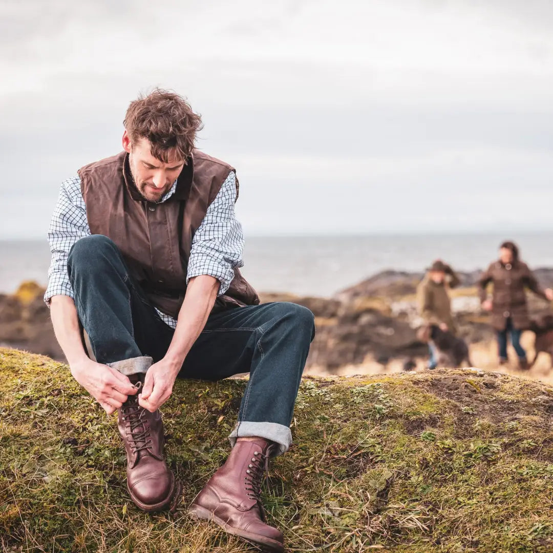 Man on mossy rock tying boot laces wearing Fife Lomond leather waistcoat