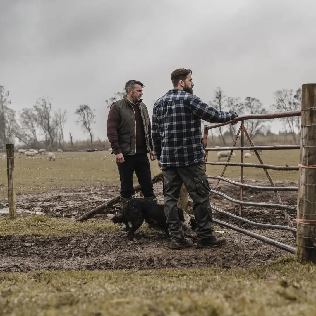 Two men and a dog by a gate wearing a Fife padded waxed waistcoat in a field