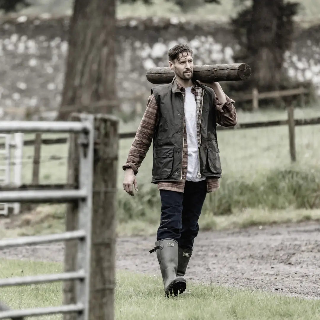 Man in a Hoggs Of Fife Padded Waxed Waistcoat carrying a log on a rural path