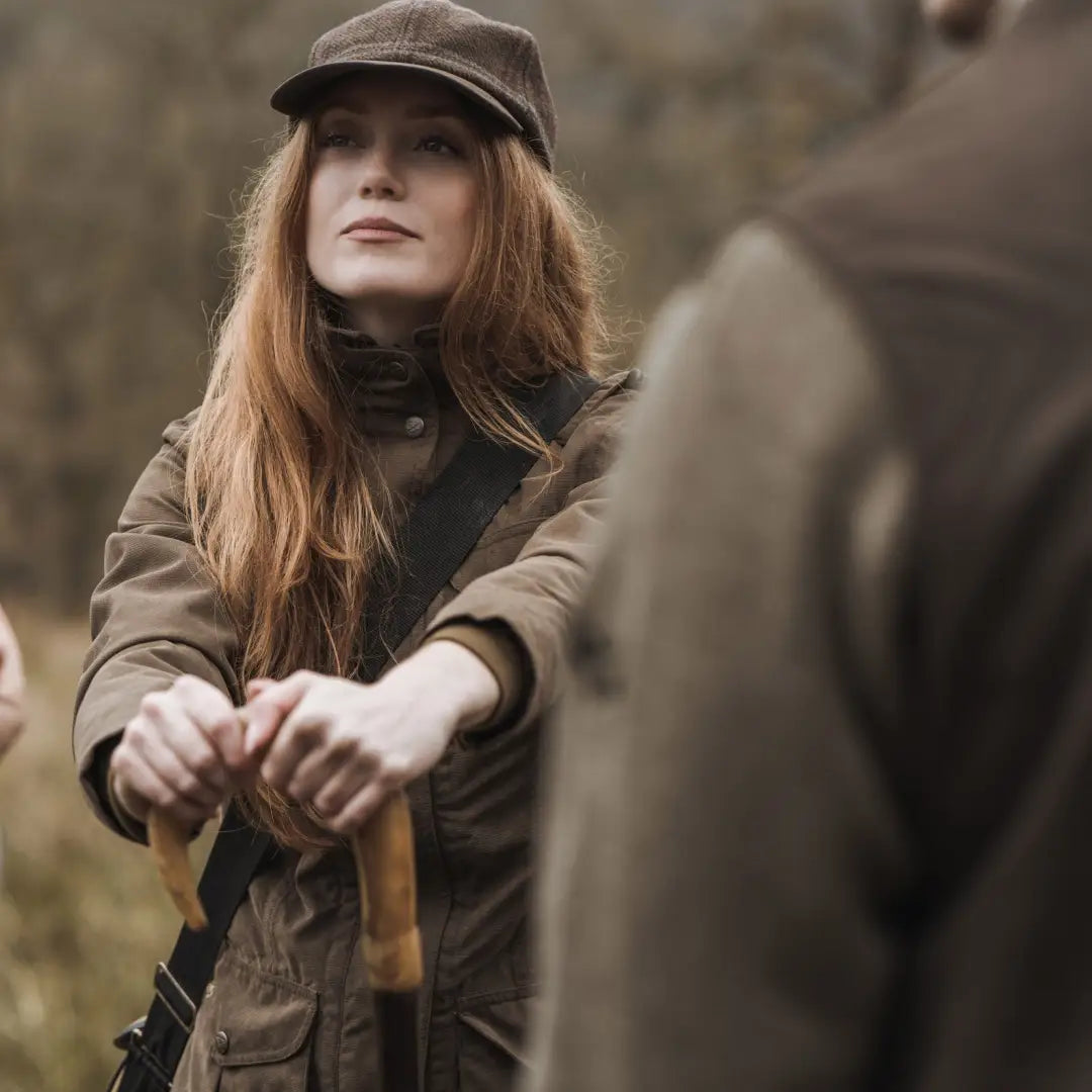 Woman with long red hair in a cap and brown coat wearing a ladies hunting jacket outdoors