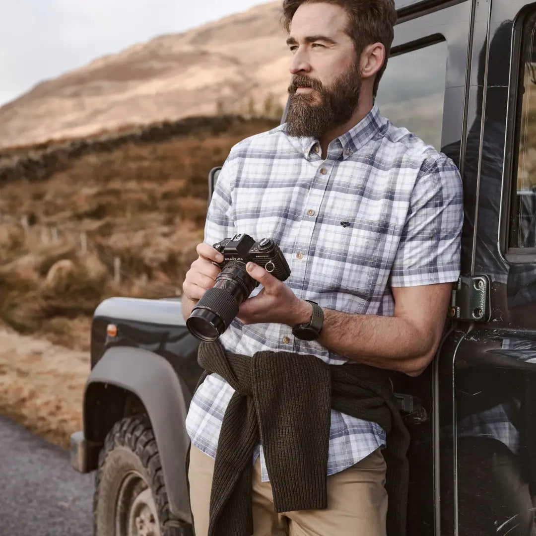 Bearded man in plaid holding binoculars near vehicle in a Fife Tresness short sleeve shirt