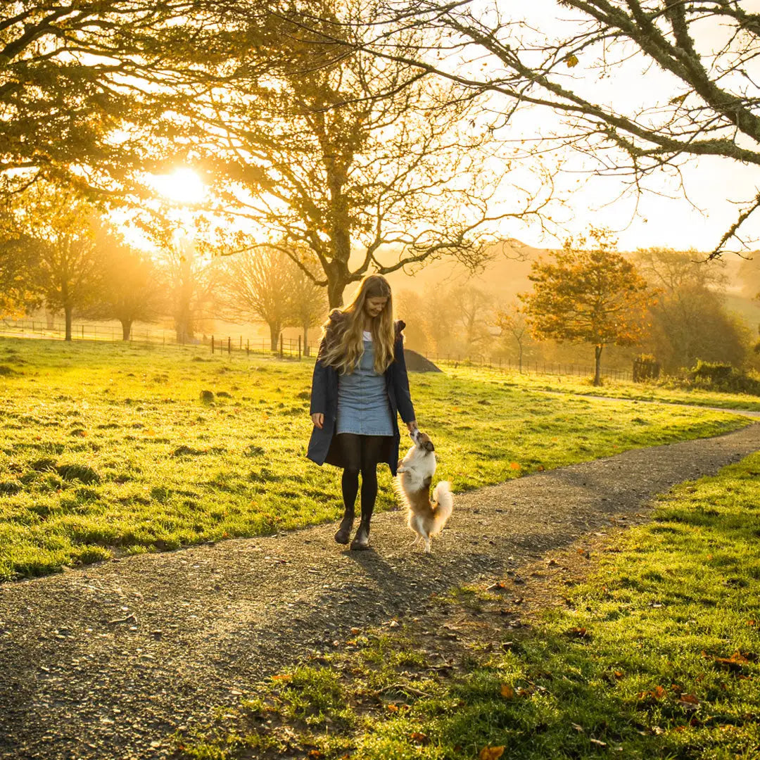 Woman in a Crofter Parka walking a dog on a sunset path, perfect for country clothing