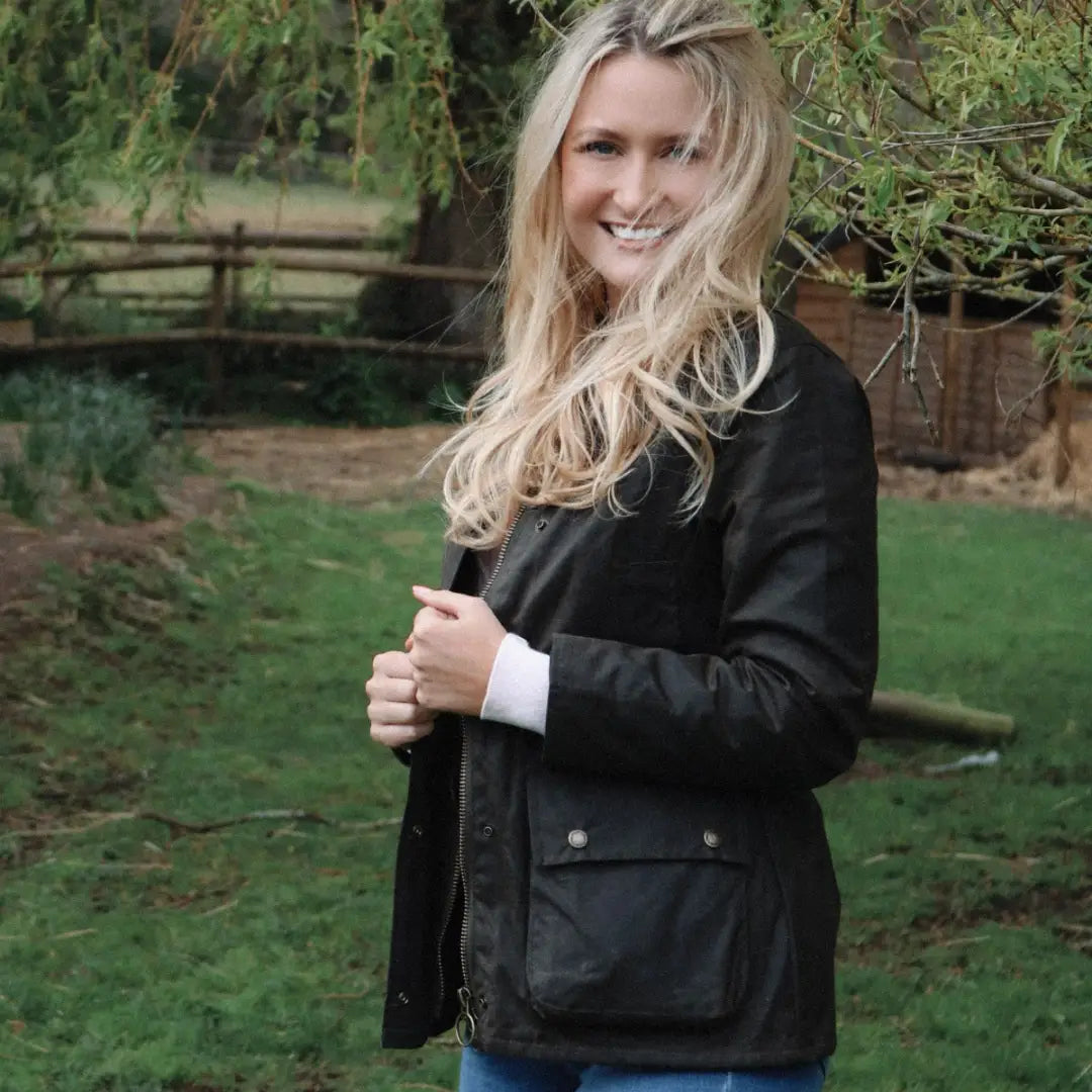 Woman with long blonde hair in a black antique wax jacket smiling outdoors