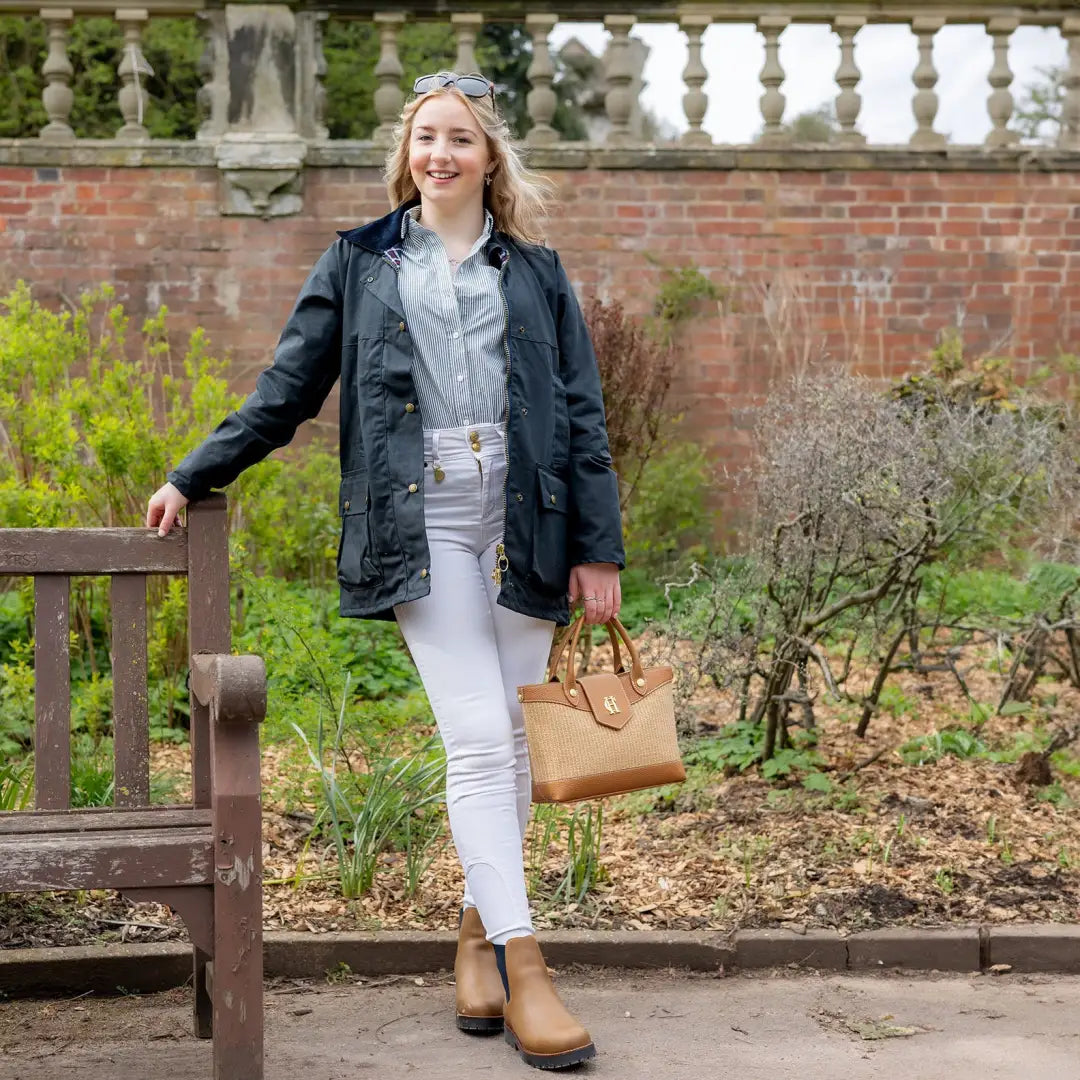 Woman in a navy blue padded wax jacket and white pants by a garden bench