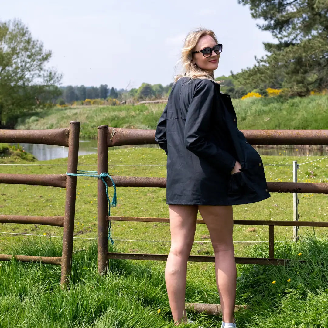 Woman in sunglasses and dark jacket by fence, showcasing Ladies Traditional Wax Jacket