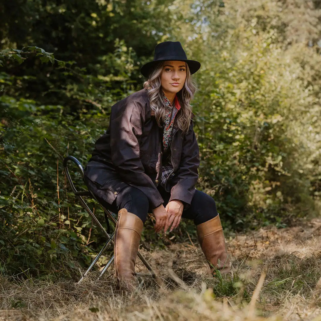 Woman in a traditional wax jacket, hat, and boots relaxing in a grassy outdoor spot