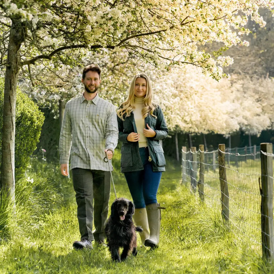 Couple in a traditional wax jacket walking a black dog under cherry blossoms