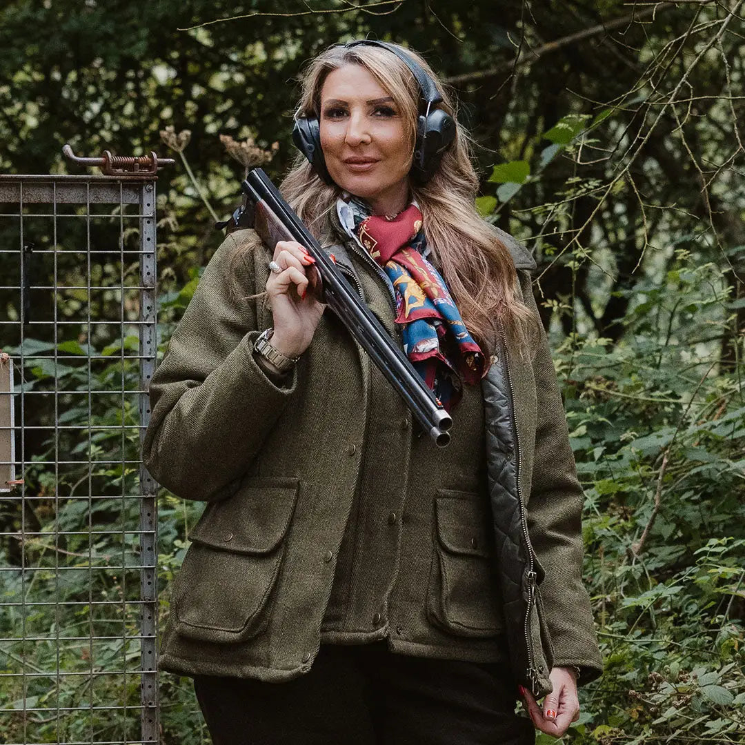 Woman in a ladies tweed jacket holding a shotgun with ear protection on