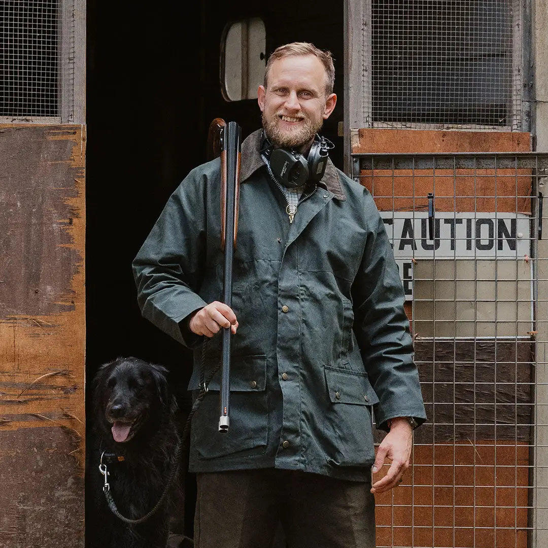 Bearded man in a green traditional wax jacket with a shovel and black dog