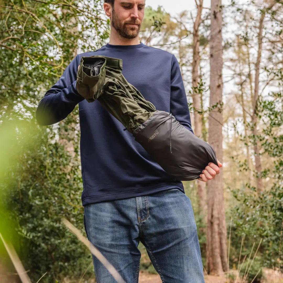 Person in a blue sweater holds a dark green sleeping bag with Musto Fenland Primaloft Jacket