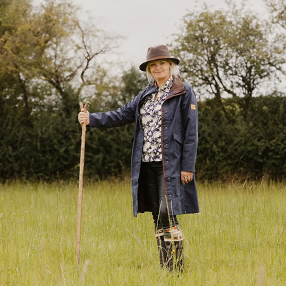 Woman in navy Forest Alice Waterproof Coat with a brown hat in a grassy field