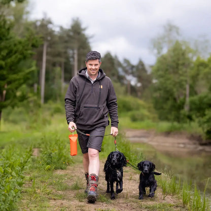 Man in Forest Heathland Hoodie walks two black dogs on a dirt path, holding water