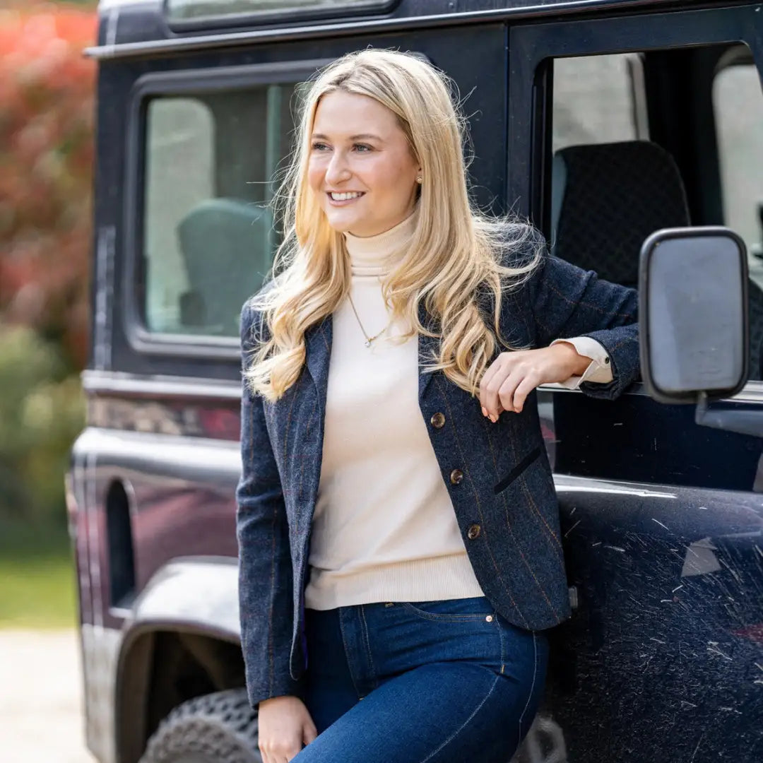 Smiling blonde woman in a white turtleneck and navy Forest Polly Tweed Blazer by a dark car