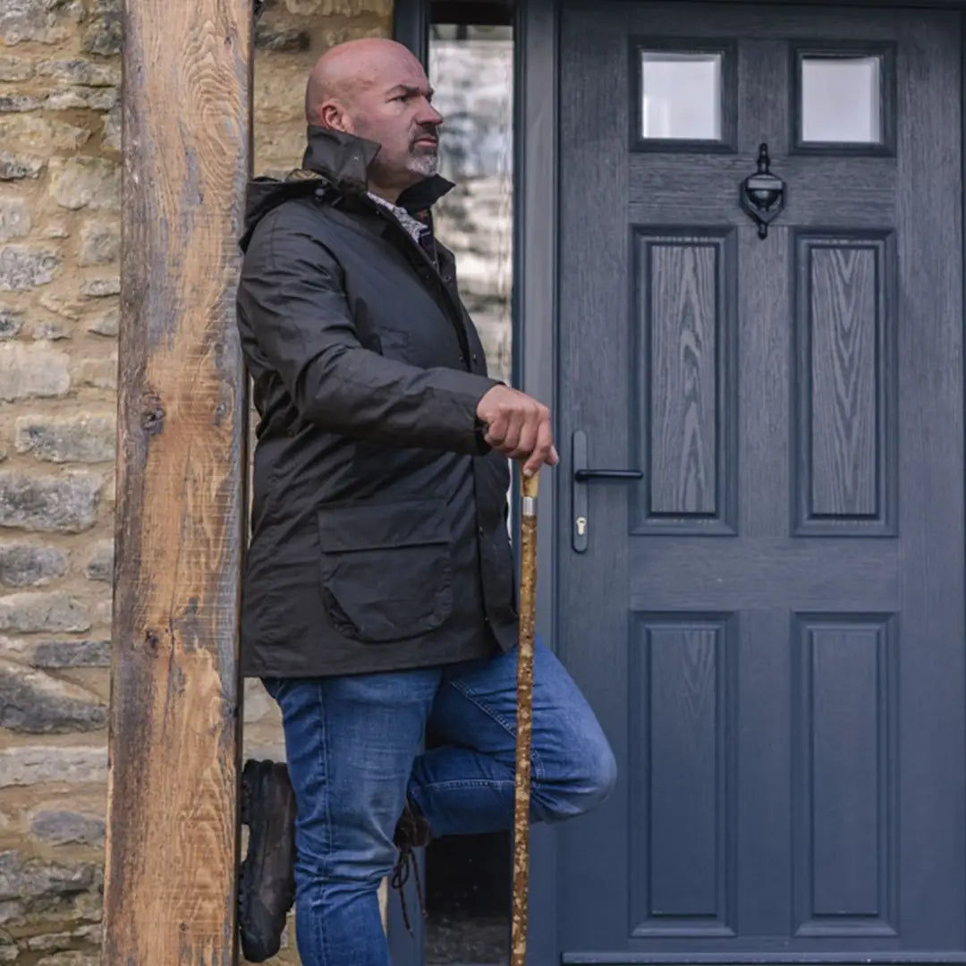 Bald man in a dark Woodsman Wax Jacket leaning against a doorframe with a walking stick