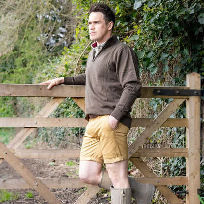 Man in brown zip neck jumper and yellow shorts leaning on a wooden fence