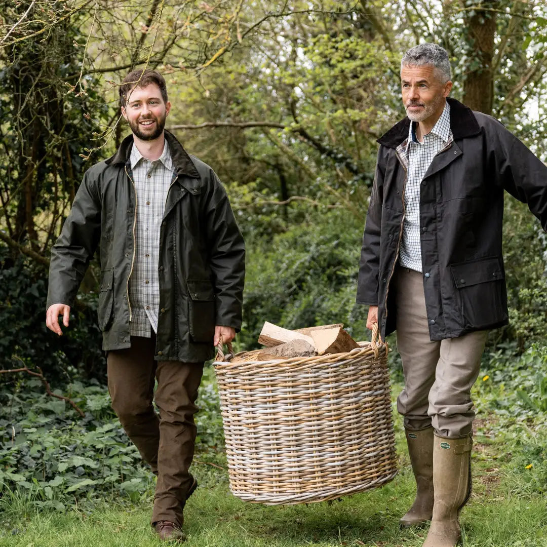 Two men in a Padded Wax Jacket carrying a wicker basket outdoors in the forest