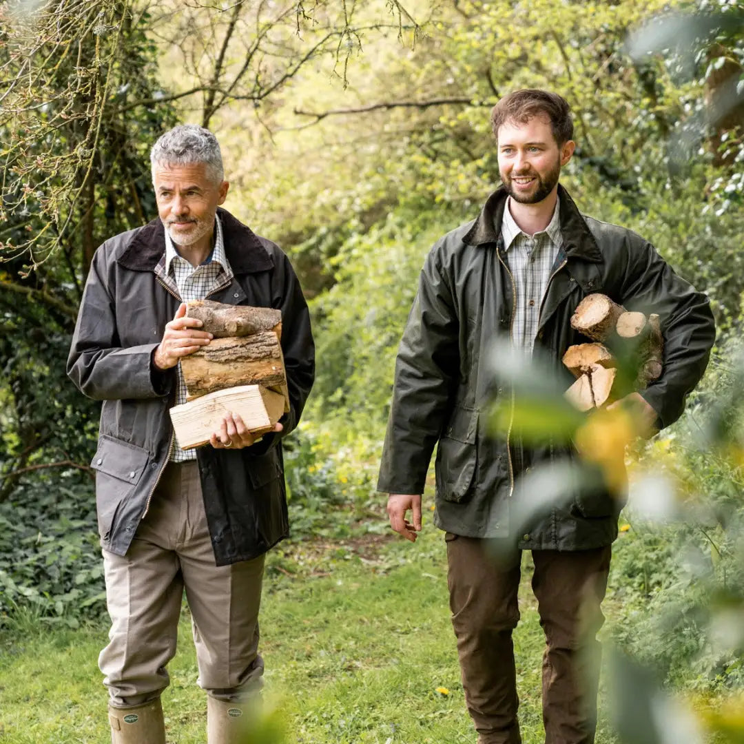 Two men in a padded wax jacket carry firewood outside in the woods