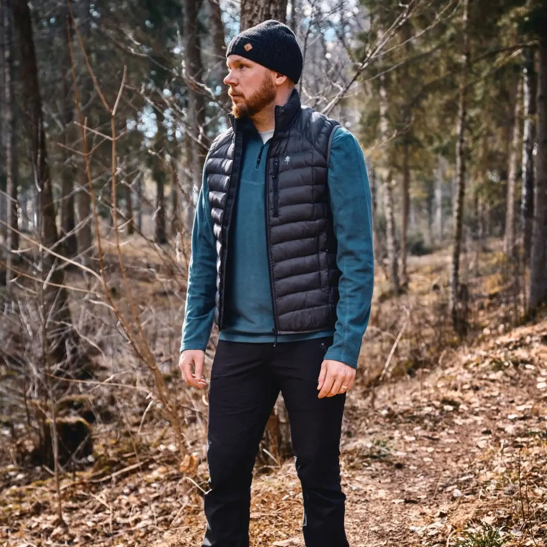 Man in a black puffer vest over a teal sweater in the woods with Pinewood Tiveden Fleece