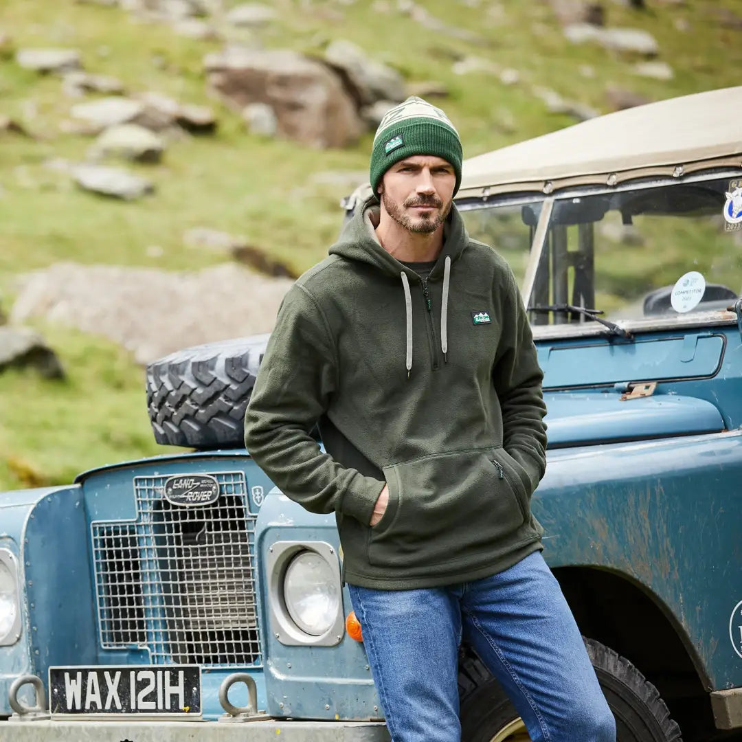 Man in a green hoodie by a blue Land Rover, showcasing the Ridgeline Ballistic Hoodie offers superior warmth