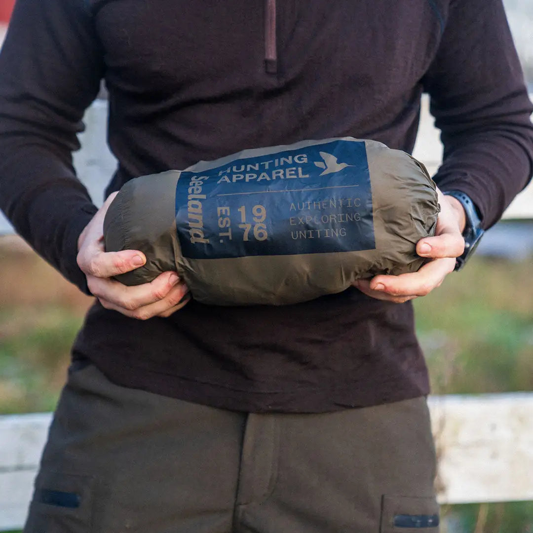 Rolled-up olive green sleeping bag next to Seeland Fahrenheit Jacket in outdoor setting