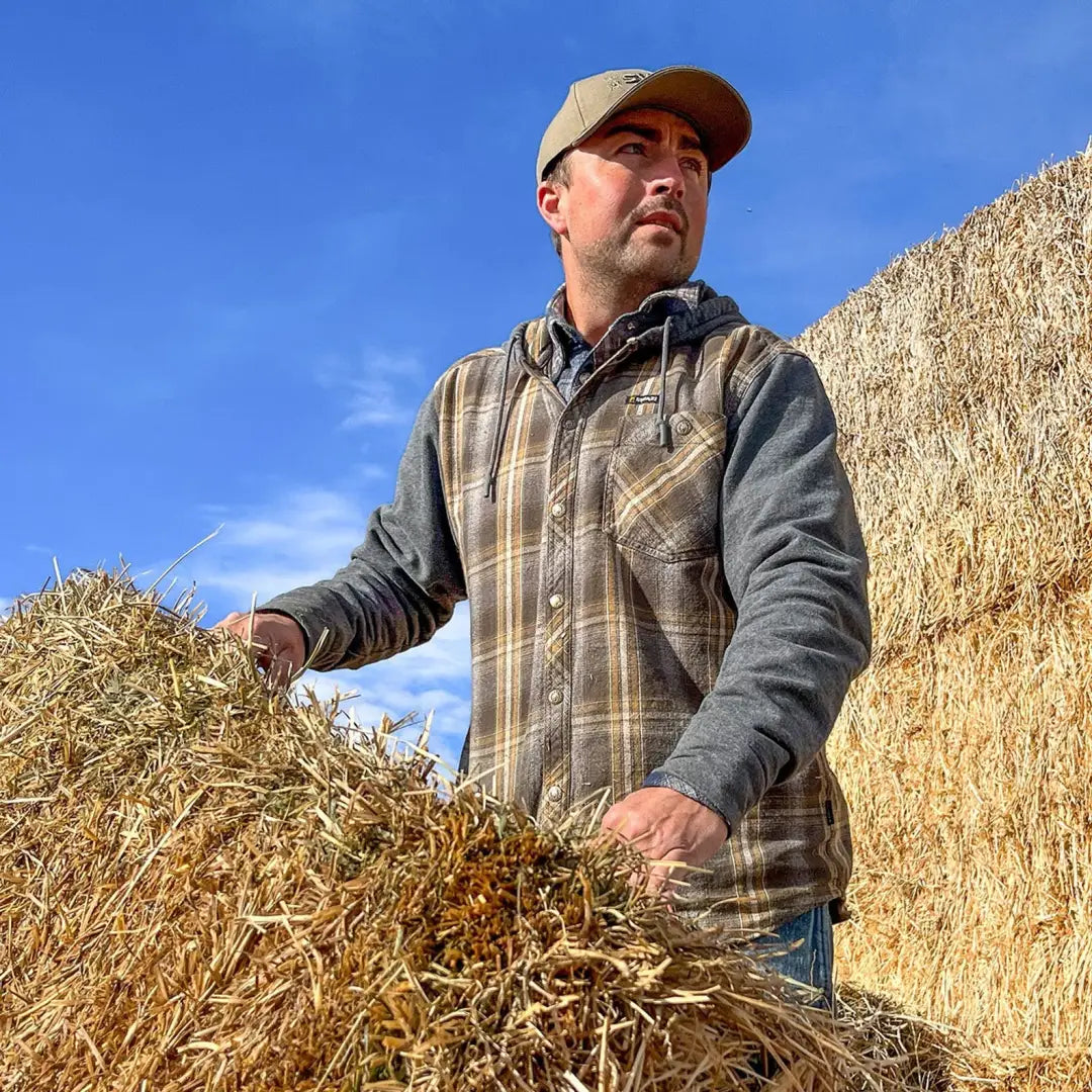 Man in a plaid vest and cap stands by hay bales, showcasing the Swazi Apprentice Hooded Shirt