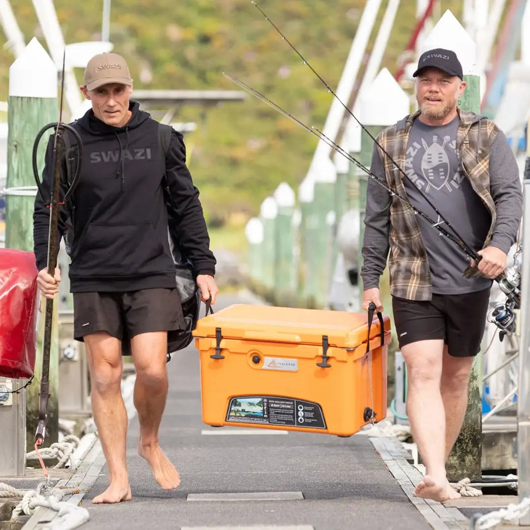 Two men in Swazi Legend Hoodies carry an orange cooler and fishing rods outdoors