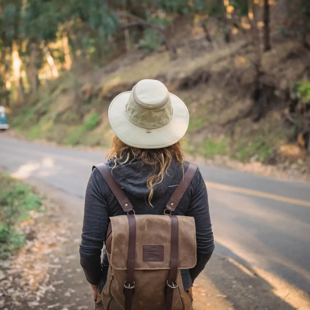 Person in a Tilley LTM6 Airflo Hat and brown leather backpack on a forest road