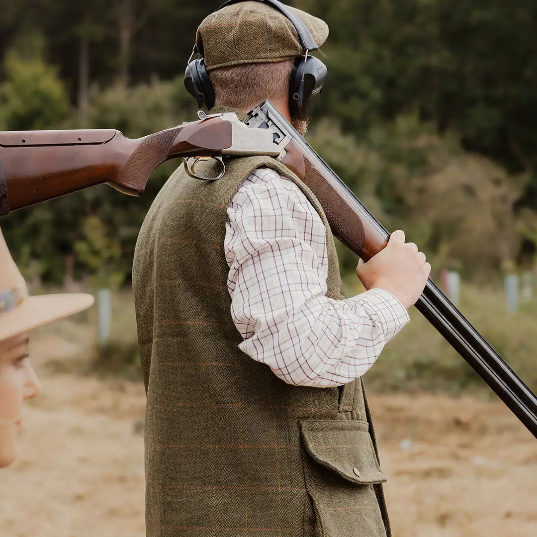 Person in hunting clothes and hearing protection holding a shotgun with a tweed gilet