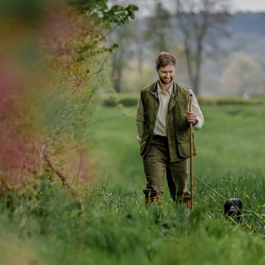 Man in a tweed gilet strolls through a grassy field with a dog and walking stick