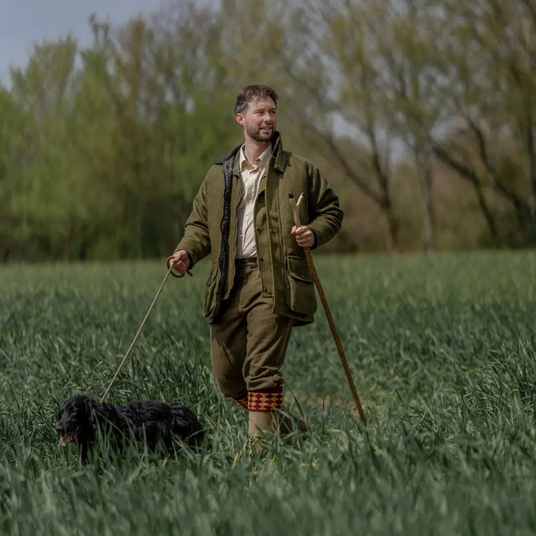 Man wearing a tweed shooting jacket walking a black dog in a grassy field